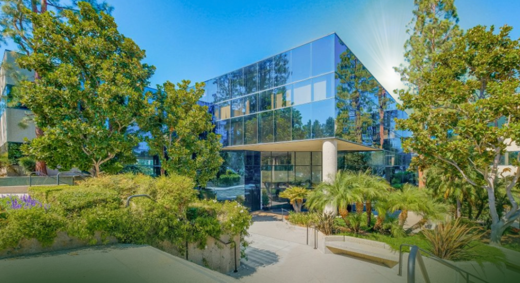 A modern glass office building surrounded by lush greenery and tall trees, with sunlight reflecting off the windows and a clear blue sky in the background.