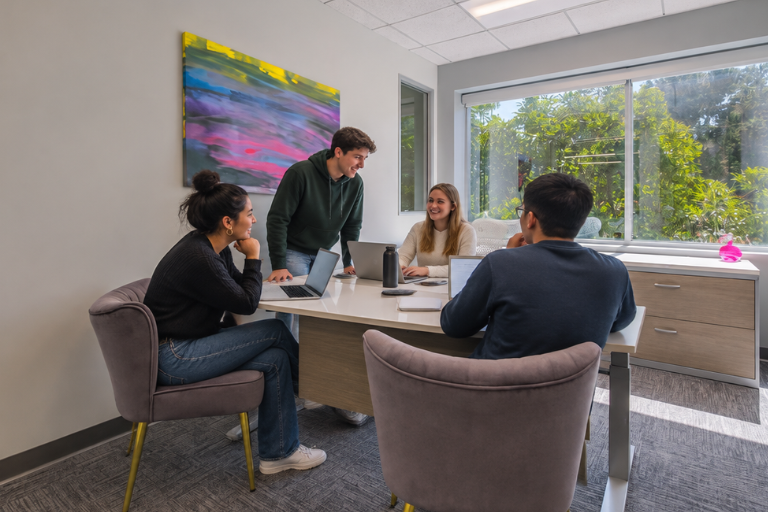 Four people sit around a desk in a modern office, talking and smiling. Laptops are open, and sunlight streams through a large window with greenery outside. A colorful abstract painting hangs on the wall.