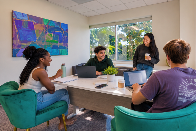 Four young adults have a meeting in a bright office with laptops and notebooks. One woman stands holding coffee, while the others sit, talking and smiling. A colorful painting and a window with greenery are visible.