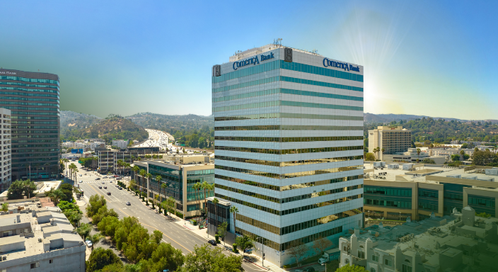 A tall office building with Comerica Bank signage stands beside a busy road, surrounded by other buildings and hills under a clear blue sky with sunlight.