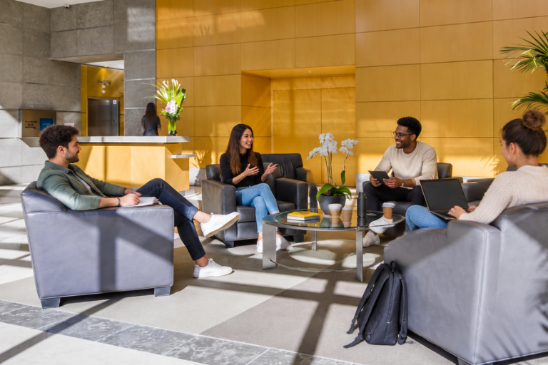 Four people sit in armchairs around a glass coffee table in a modern office lobby, talking and smiling. Laptops and notebooks are on the table, and a backpack is on the floor nearby.