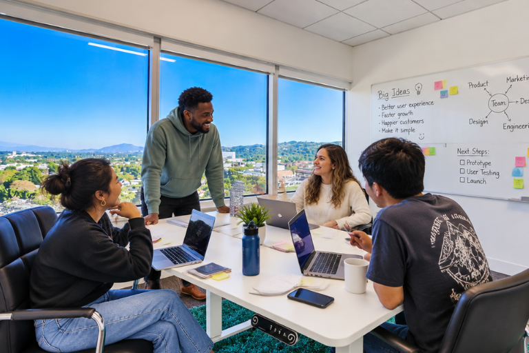 Four people sit around a white table with laptops and notebooks, having a meeting in a bright office with large windows and a whiteboard filled with notes. A cityscape and hills are visible outside.