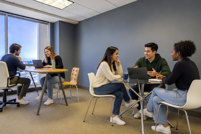 Two small groups of young adults sit at round tables in a modern office, talking and working on laptops. Large windows provide natural light, and everyone appears engaged in friendly conversation.