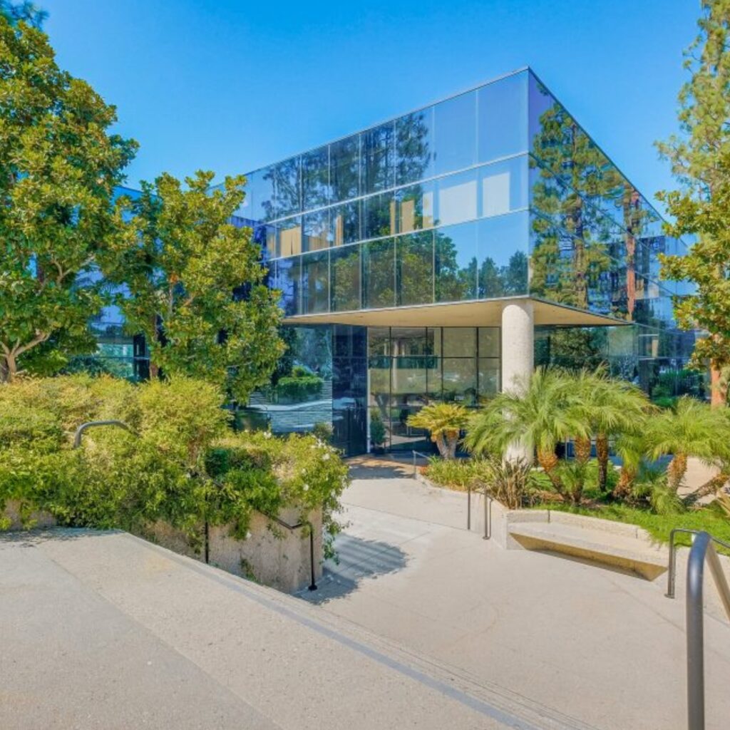 Modern glass office building surrounded by trees and greenery, with sunlight reflecting on the windows and a concrete path leading to the entrance.