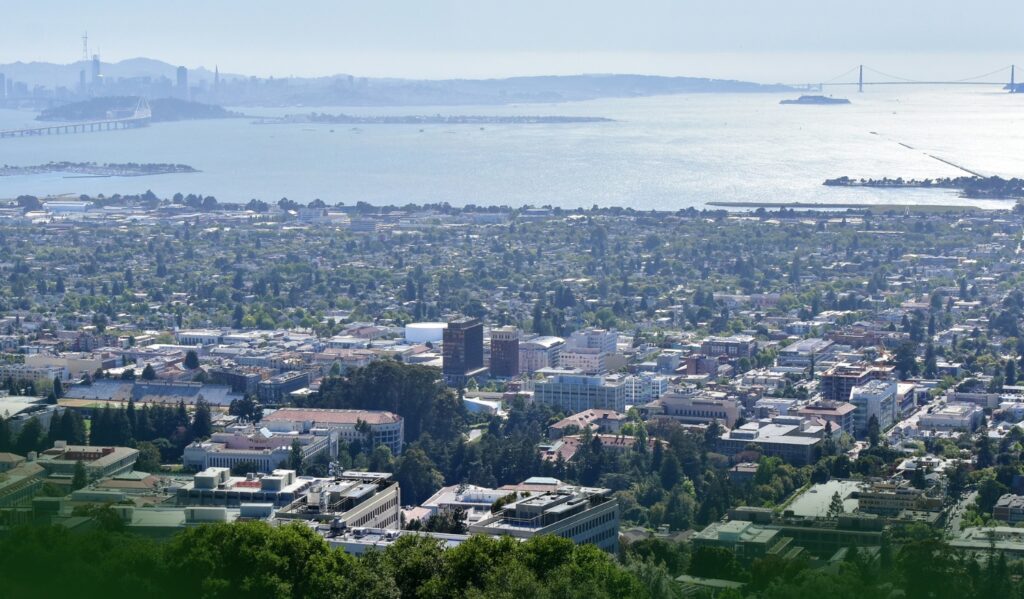 Aerial view of a city by the bay, with buildings in the foreground, water in the middle, and a distant bridge and hazy city skyline in the background.