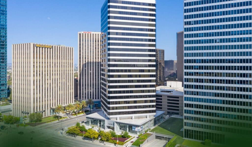 A group of tall office buildings in a city, with a Wells Fargo building on the left, palm trees, landscaped areas, and cars driving on city streets under a clear blue sky.
