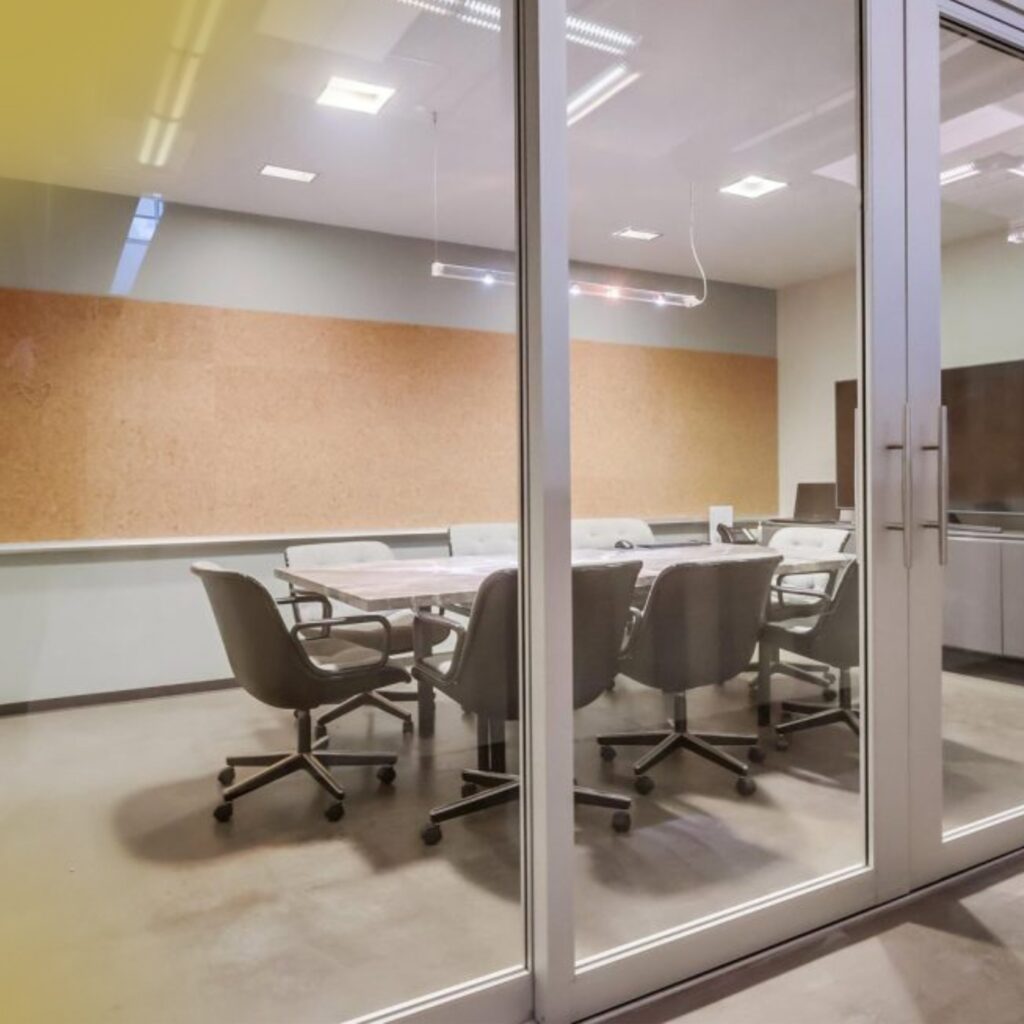 A modern conference room with a glass door, gray office chairs around a rectangular table, a corkboard wall, and overhead lighting.