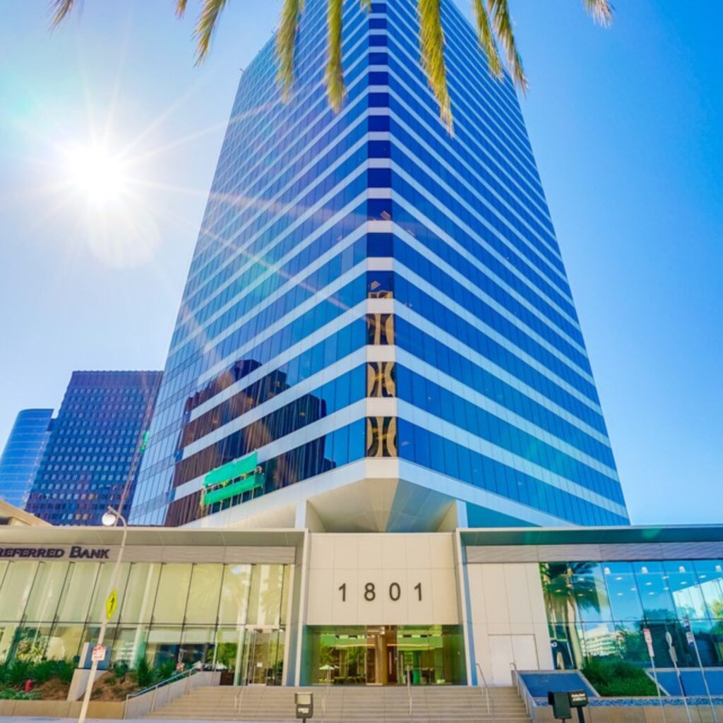 A tall glass office building with the number 1801 at the entrance, palm trees, and sunlight shining in the corner against a clear blue sky.