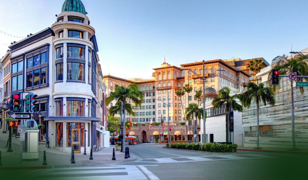 Sunny city street with palm trees, a unique rounded building on the corner, luxury shops, colorful traffic lights, and a crosswalk under a clear blue sky.