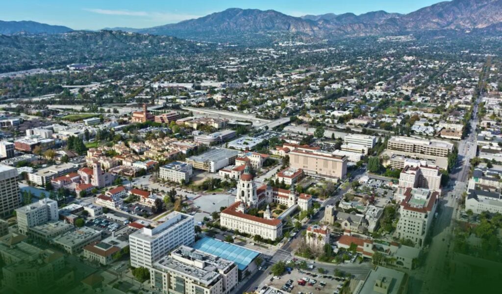 Aerial view of a city with a mix of low-rise buildings, tree-lined streets, and distant mountains under a blue sky.