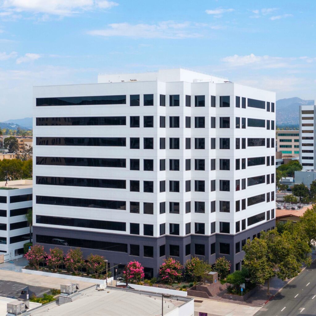 A modern, multi-story office building with white and black horizontal stripes, surrounded by trees and other buildings, under a partly cloudy sky.
