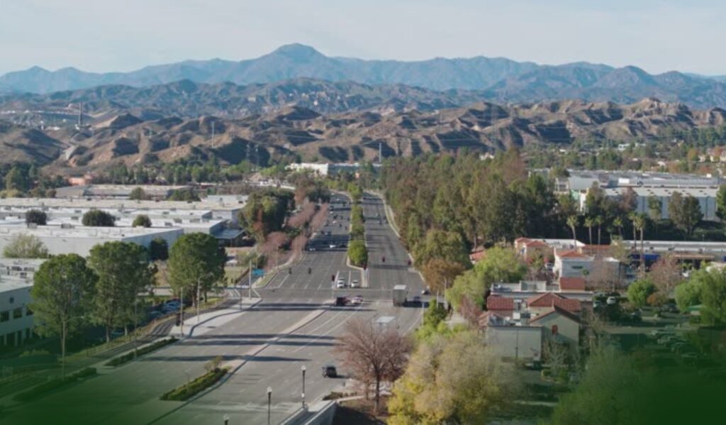 A wide street runs through a suburban area with buildings and trees, leading toward distant brown hills and mountains under a clear sky.