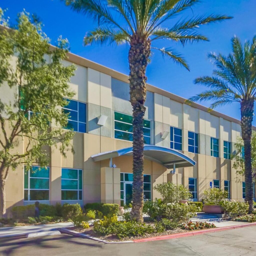 Modern two-story office building with large windows, tan and gray exterior, palm trees, landscaped bushes, and a curved metal entrance awning under a bright blue sky.
