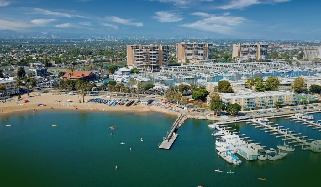 A marina with boats docked, a sandy beach, and people kayaking. Tall buildings and a city skyline are visible in the background under a partly cloudy sky.