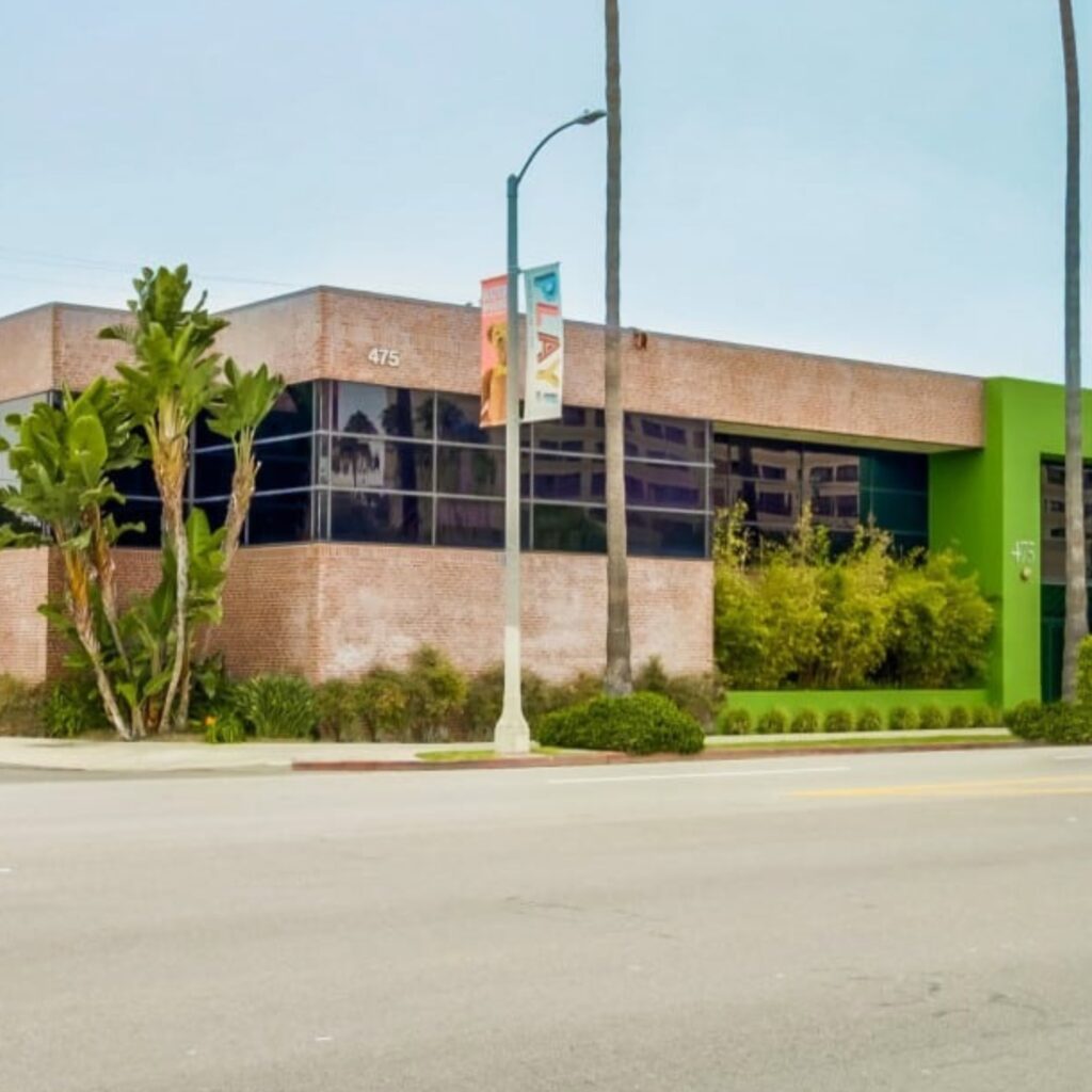 A modern, low-rise brick office building with green trim, tall windows, and surrounding greenery sits on a quiet street under a cloudy sky.