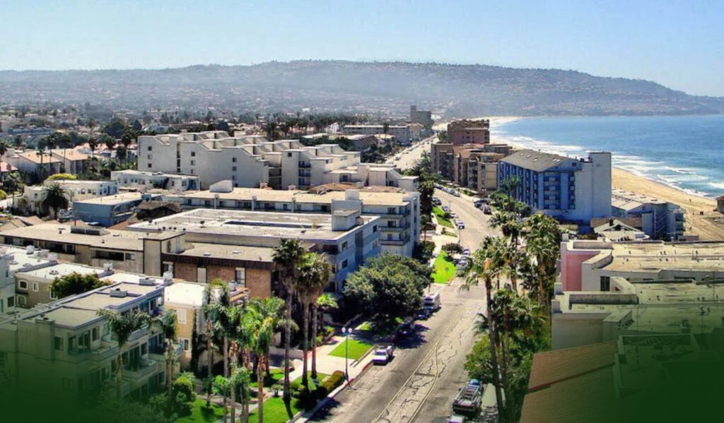 A coastal cityscape with palm trees, mid-rise buildings, and a road running parallel to a sandy beach and the ocean, with hills in the background under a clear sky.