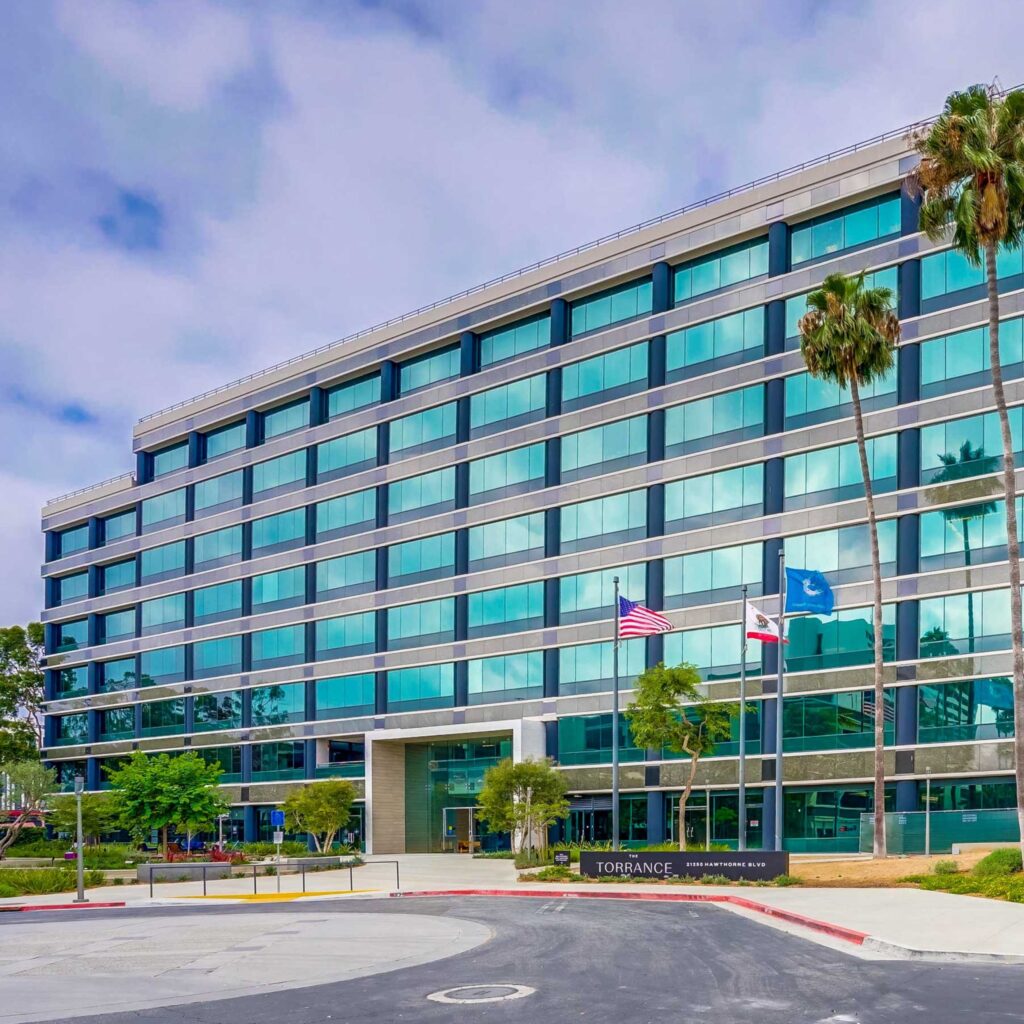 Modern glass building with six stories, palm trees in front, and three flags flying by the entrance. The sign reads Torrance. The sky is partly cloudy.
