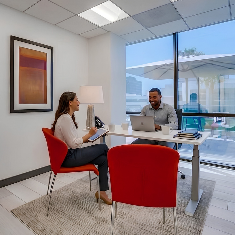 Two people sit across from each other at a desk in a modern office, smiling and talking. Large windows let in natural light, and vibrant red chairs add color to the bright, tidy space.