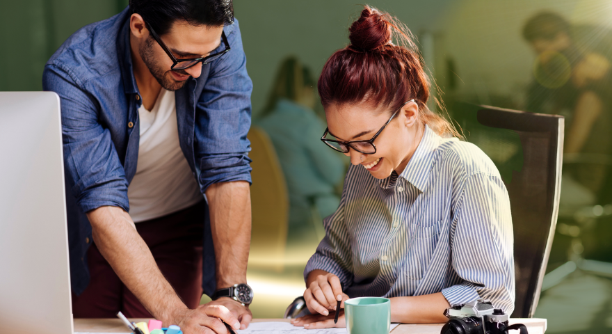 Two people in an office smiling and collaborating at a desk, looking at a document. There is a computer, coffee mug, and camera on the desk. The setting is bright and professional.