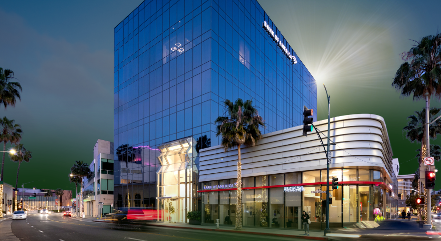 A modern glass building with palm trees in front sits on a street corner at dusk. The lower level has a Bank of America branch, and traffic lights are visible at the intersection.