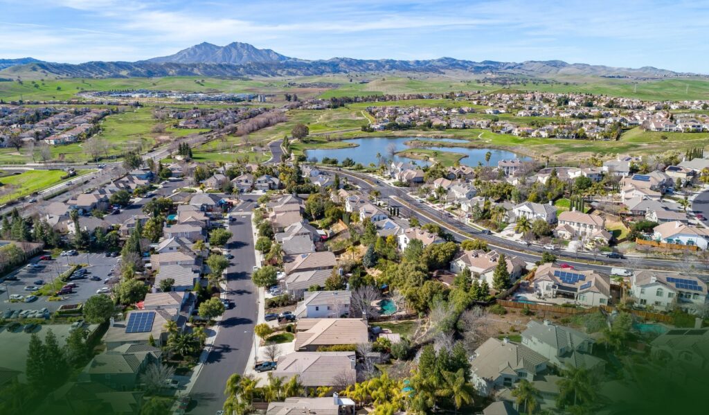 Aerial view of a suburban neighborhood with houses, roads, green lawns, a small lake, and distant mountains under a blue sky.