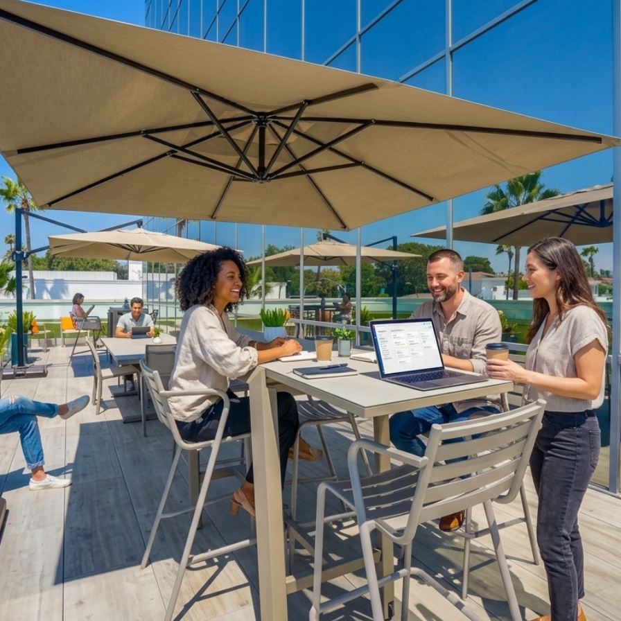 Three people work at a high table on a sunny outdoor terrace shaded by large umbrellas. A woman sits, a man sits with a laptop, and another woman stands nearby with a coffee cup. They appear to be in a professional setting near a glass building.