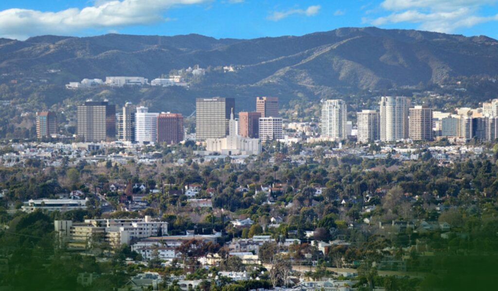 City skyline with tall buildings in front of tree-filled neighborhoods, set against a backdrop of mountains and a partly cloudy sky.