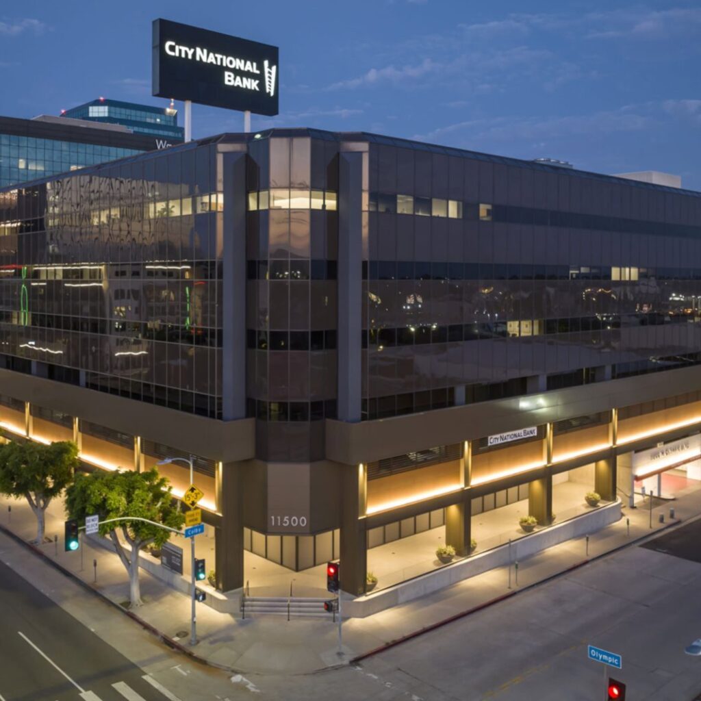 A modern office building at dusk with City National Bank signage on the roof and street signs for Olympic Blvd and Braddock Dr at the corner. The building is well-lit and features reflective glass windows.