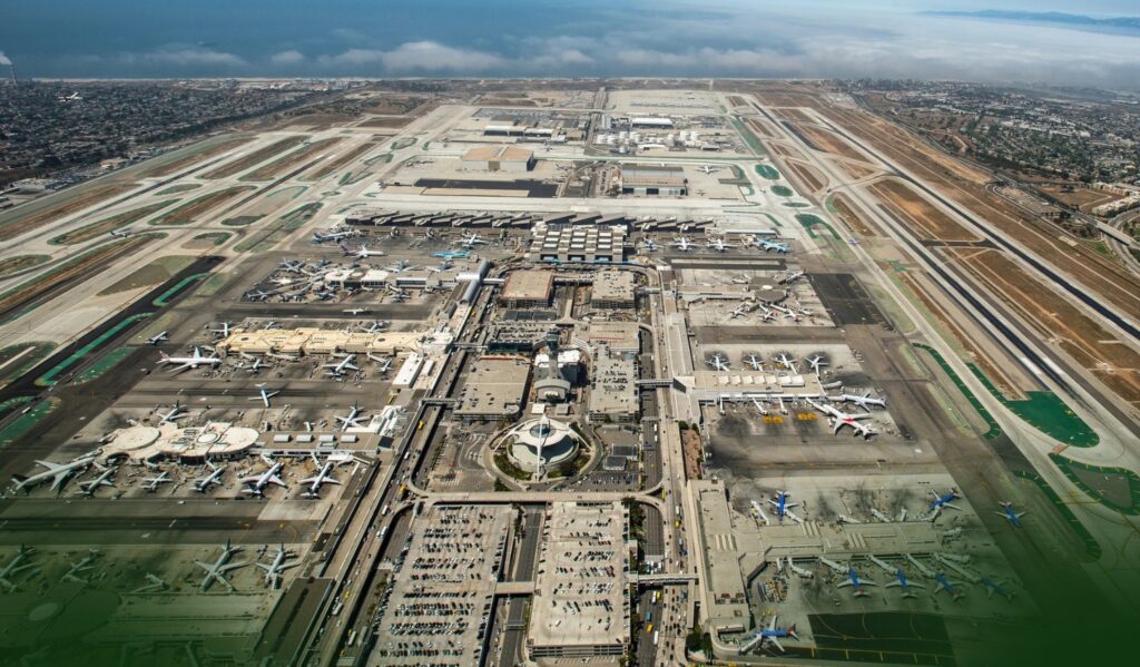 Aerial view of a large airport with multiple runways, taxiways, terminals, and airplanes parked at gates; cityscape and ocean in the background under a cloudy sky.