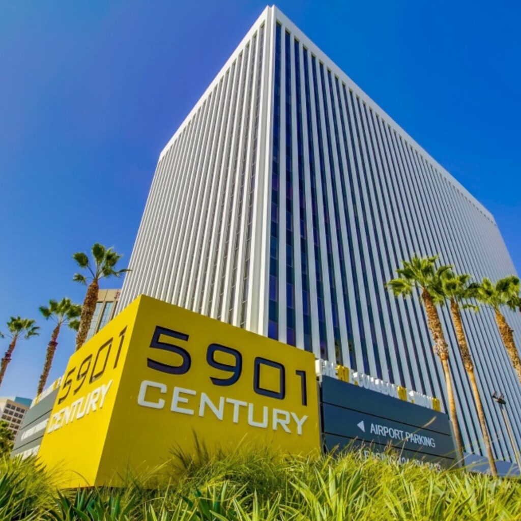 A tall office building with vertical stripes stands behind a yellow sign that reads 5901 Century. Palm trees line the sidewalk, and the sky is clear and blue.
