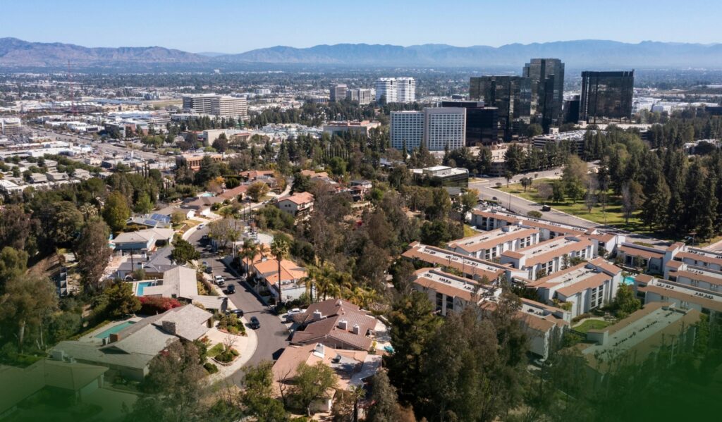 Aerial view of a suburban neighborhood with houses, trees, and nearby office buildings, set against distant mountains under a clear blue sky.