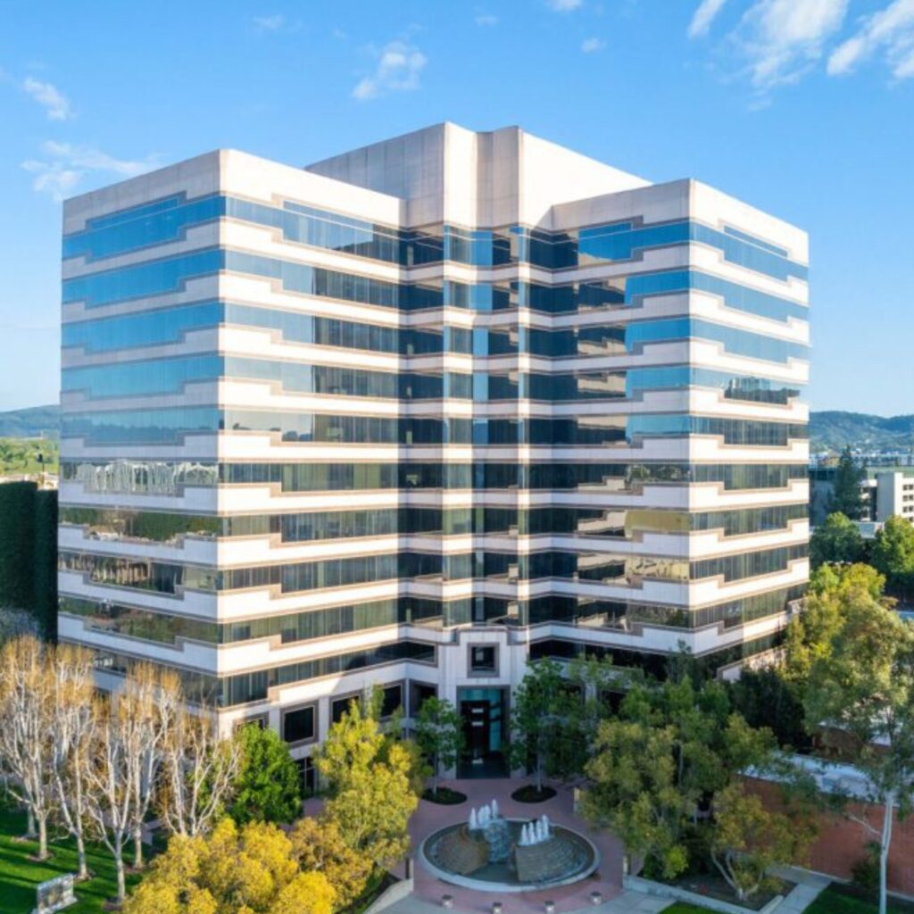 A modern, multi-story office building with reflective glass windows, surrounded by trees and landscaping under a blue sky with clouds.