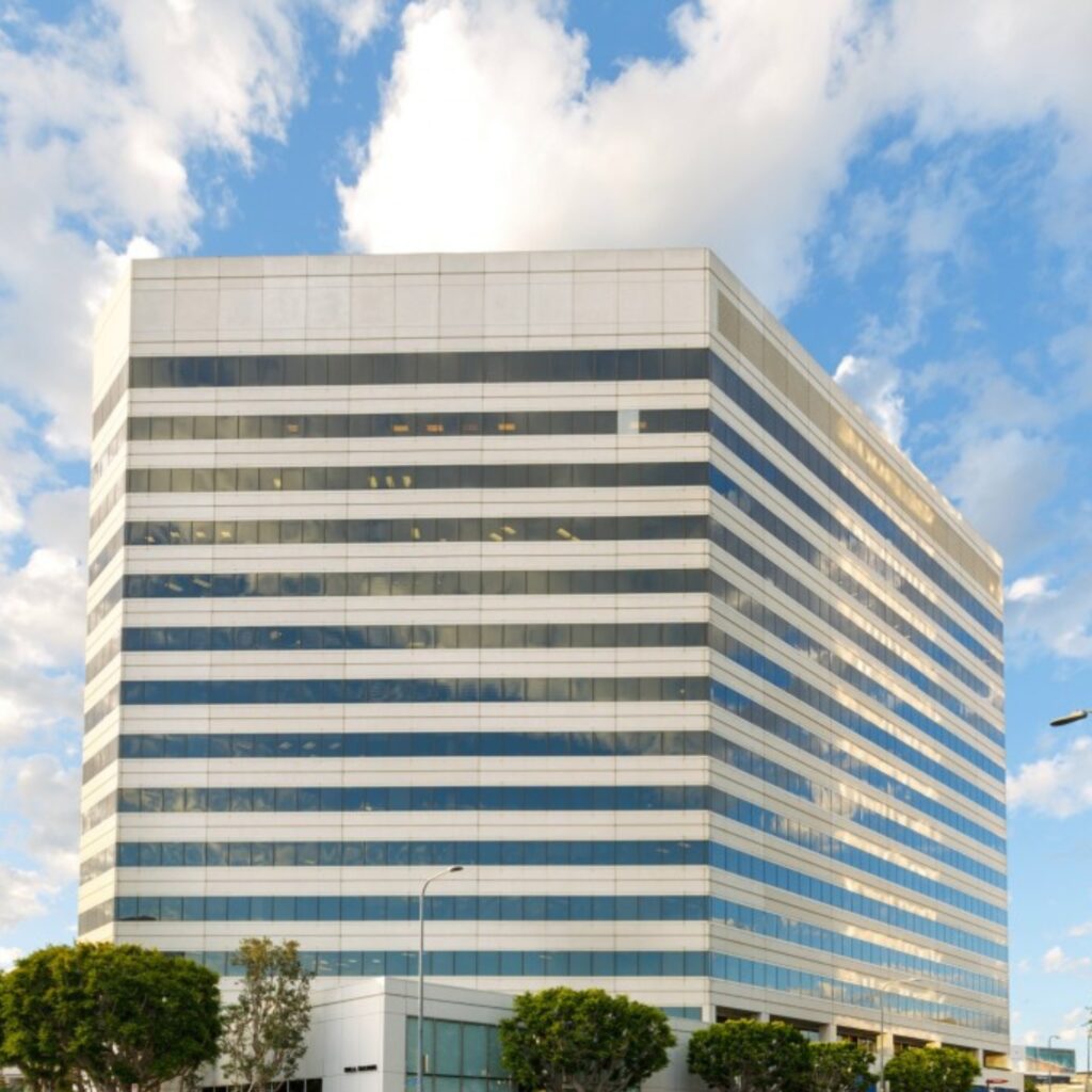 A modern, multi-story office building with reflective windows stands under a blue sky with scattered clouds, surrounded by small green trees and streetlights.