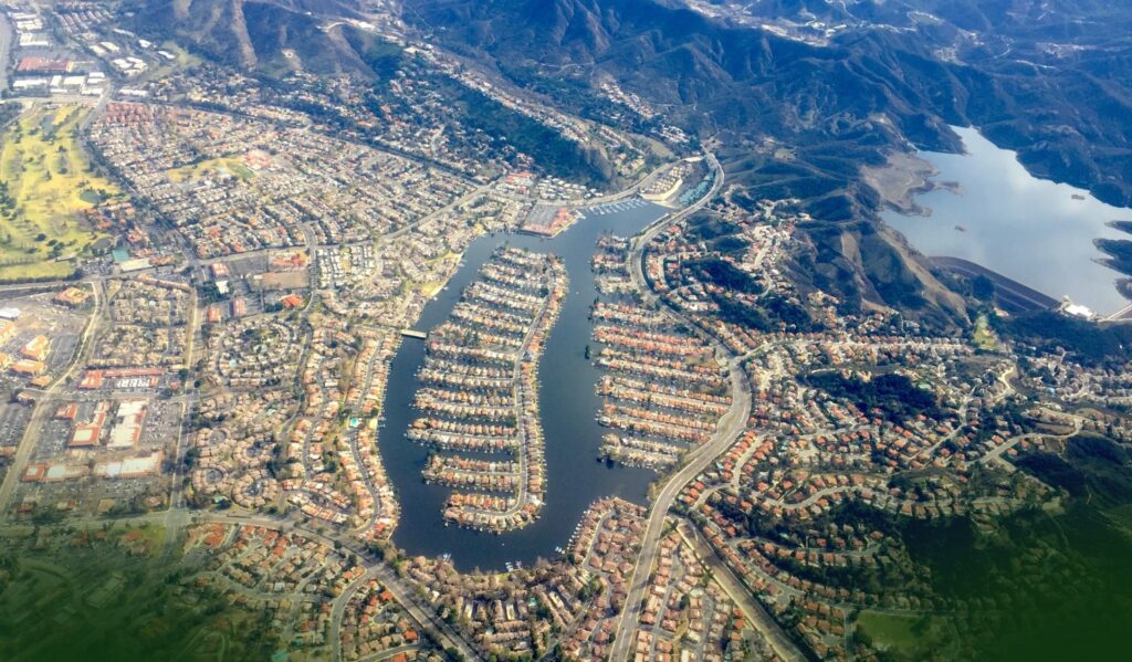 Aerial view of a suburban neighborhood with houses built along winding waterways, surrounded by mountains and a large lake in the distance.