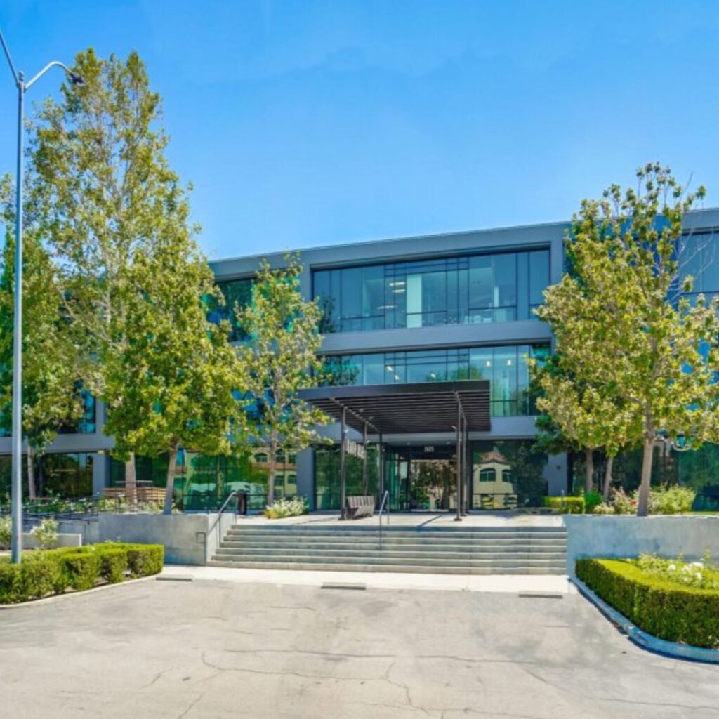 Modern office building with large windows and a shaded entrance, surrounded by green trees and shrubs. Concrete steps lead up from a mostly empty parking lot under a clear blue sky.