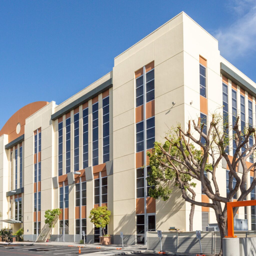 Modern four-story office building with tall vertical windows, beige walls with orange accents, and a few small trees in front. The sky is clear and blue, and there are some parking spaces visible at the bottom.