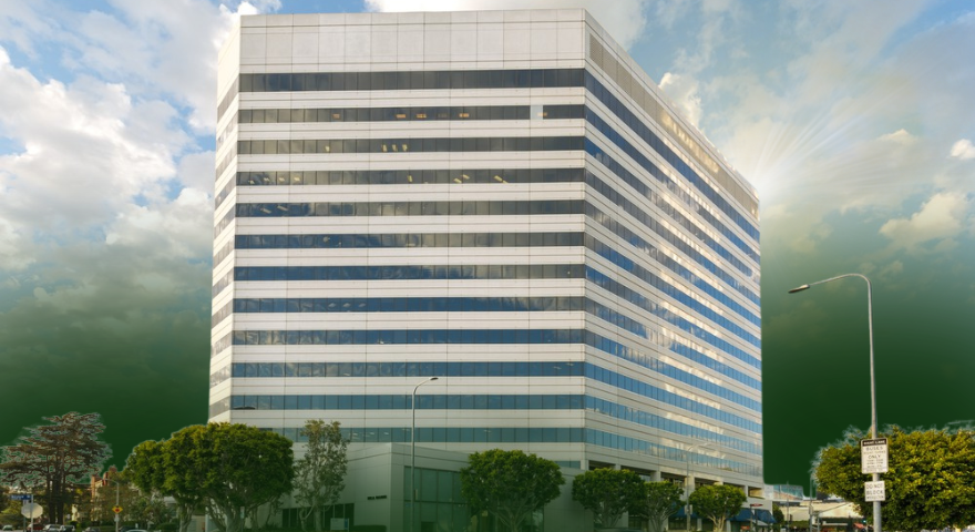 A modern, multi-story glass office building stands on a city corner, surrounded by trees and streetlights, with a partly cloudy sky and sunlight shining in the background.