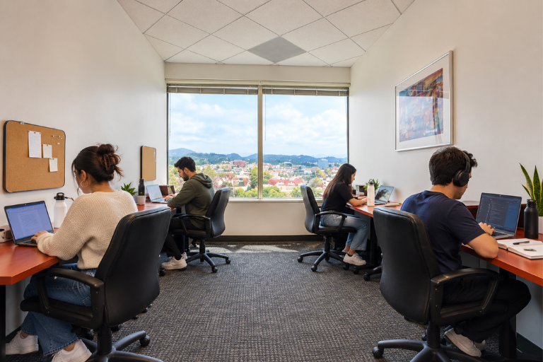 Four people work at desks in a small, bright office with a large window showing a cityscape. They use laptops, some wear headphones. The room has cork boards, plants, and a colorful framed picture on the wall.
