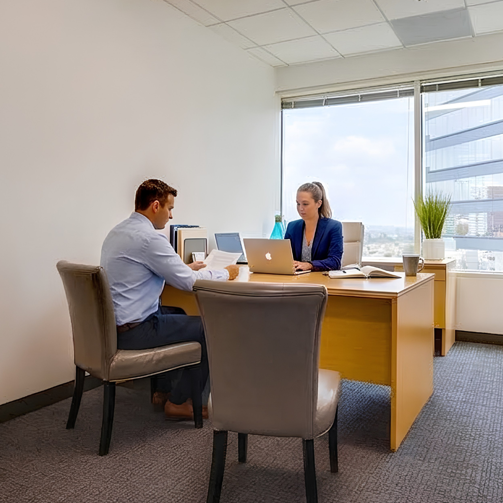 A man and a woman sit across from each other at a desk in a modern office. The woman works on a laptop, while the man looks at documents. Large windows show a cityscape outside.