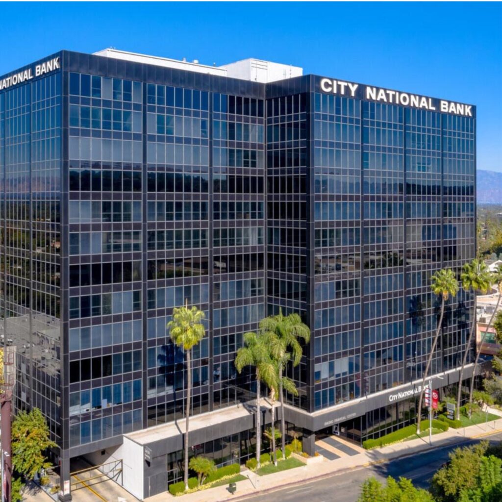 A modern glass office building with City National Bank signage, palm trees in front, and a clear sky in the background.