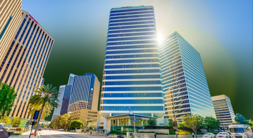 Tall modern office buildings with glass windows line a sunny city street, palm trees and greenery in front, and bright sunlight reflecting off the central tower.