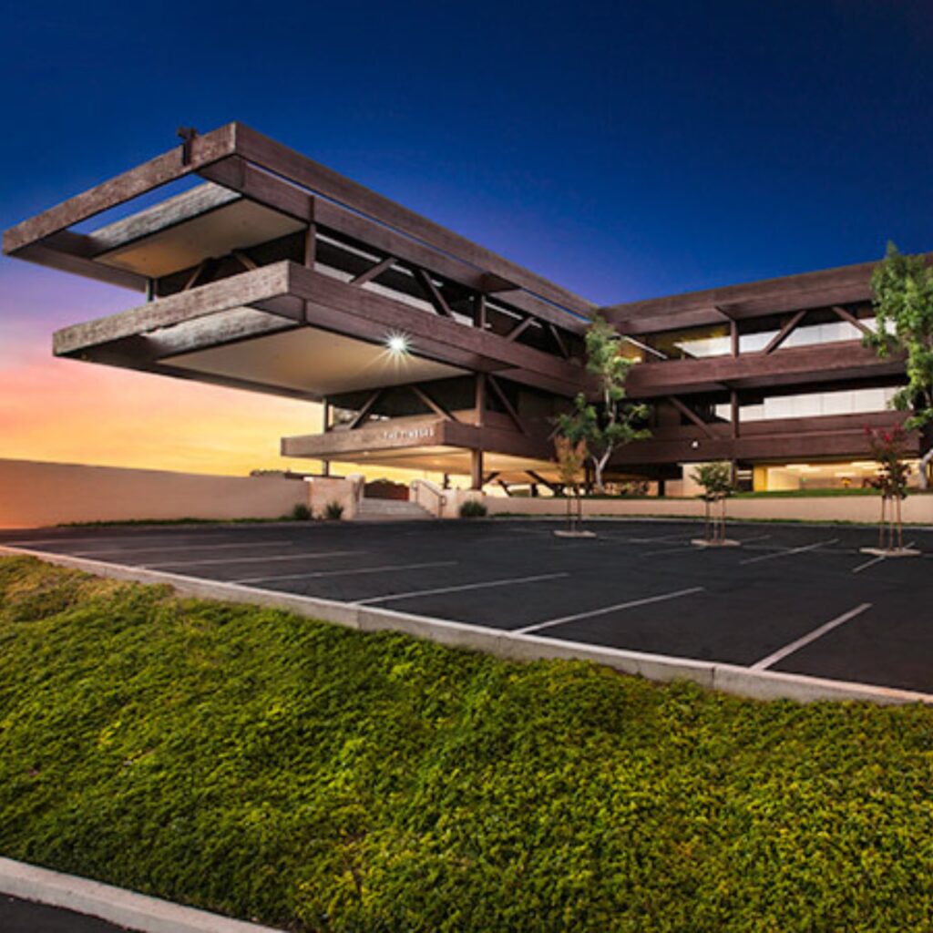 Modern three-story building with angular, cantilevered sections, large windows, and a mostly empty parking lot at dusk. Green landscaping is in the foreground under a vibrant sunset sky.