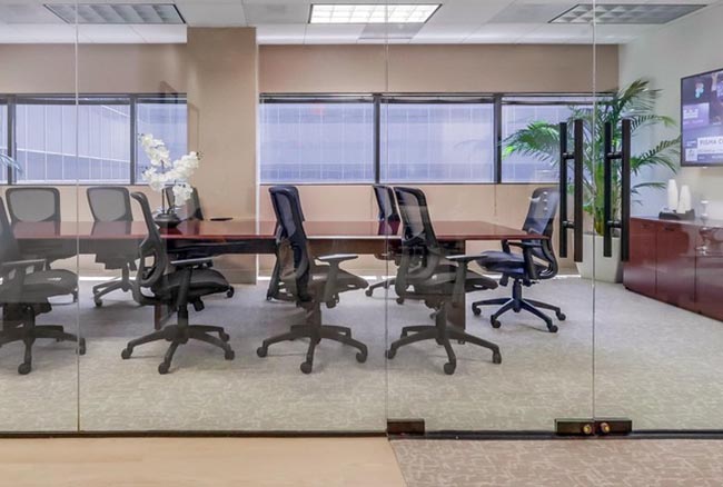 A modern conference room with a long table, six black office chairs, glass walls, large windows, a potted plant, and a sideboard with a TV monitor.