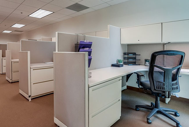 A modern office with empty cubicles, beige walls, white desks, mesh office chairs, and overhead storage, arranged in a row under fluorescent ceiling lights.