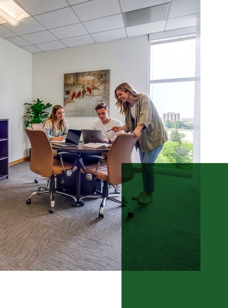 Three people collaborate at a table in a bright office with large windows, a plant, and wall art. Two are seated with laptops, while one stands, smiling and engaging with the group. A green transparent shape overlays the image.