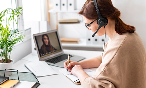 A woman wearing a headset takes notes while attending a video call on her laptop in a bright home office, with a plant and folders in the background.