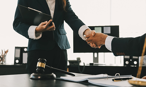 Two people in business attire shake hands in an office with legal documents, a gavel, and a clipboard on the desk, suggesting a legal or business agreement.