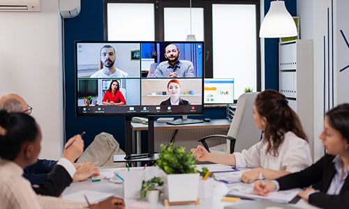 Four people sit at a conference table, participating in a video call with four others shown on a large screen in a modern office. Notepads, pens, and plants are on the table.