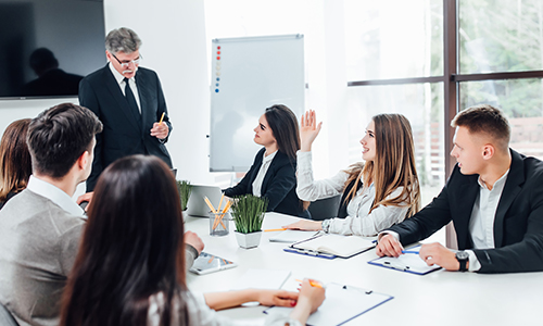 A group of six people in business attire are in a meeting room. One woman raises her hand while an older man stands and speaks. Others sit at a table with notebooks and laptops. A whiteboard and windows are in the background.