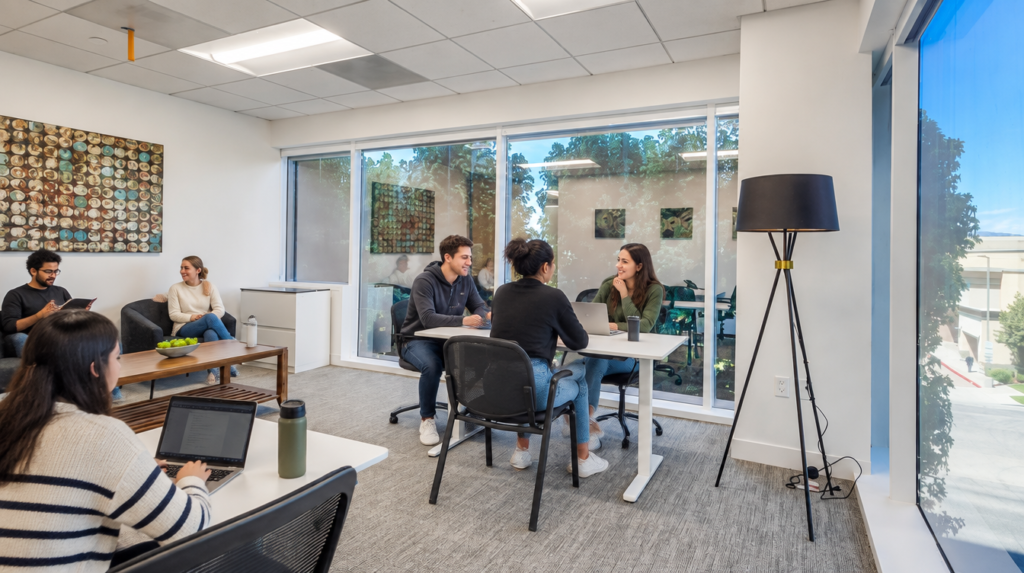 Five people are in a bright, modern office. Two pairs talk at tables, one person works on a laptop, and another reads on a couch. Large windows show greenery outside.